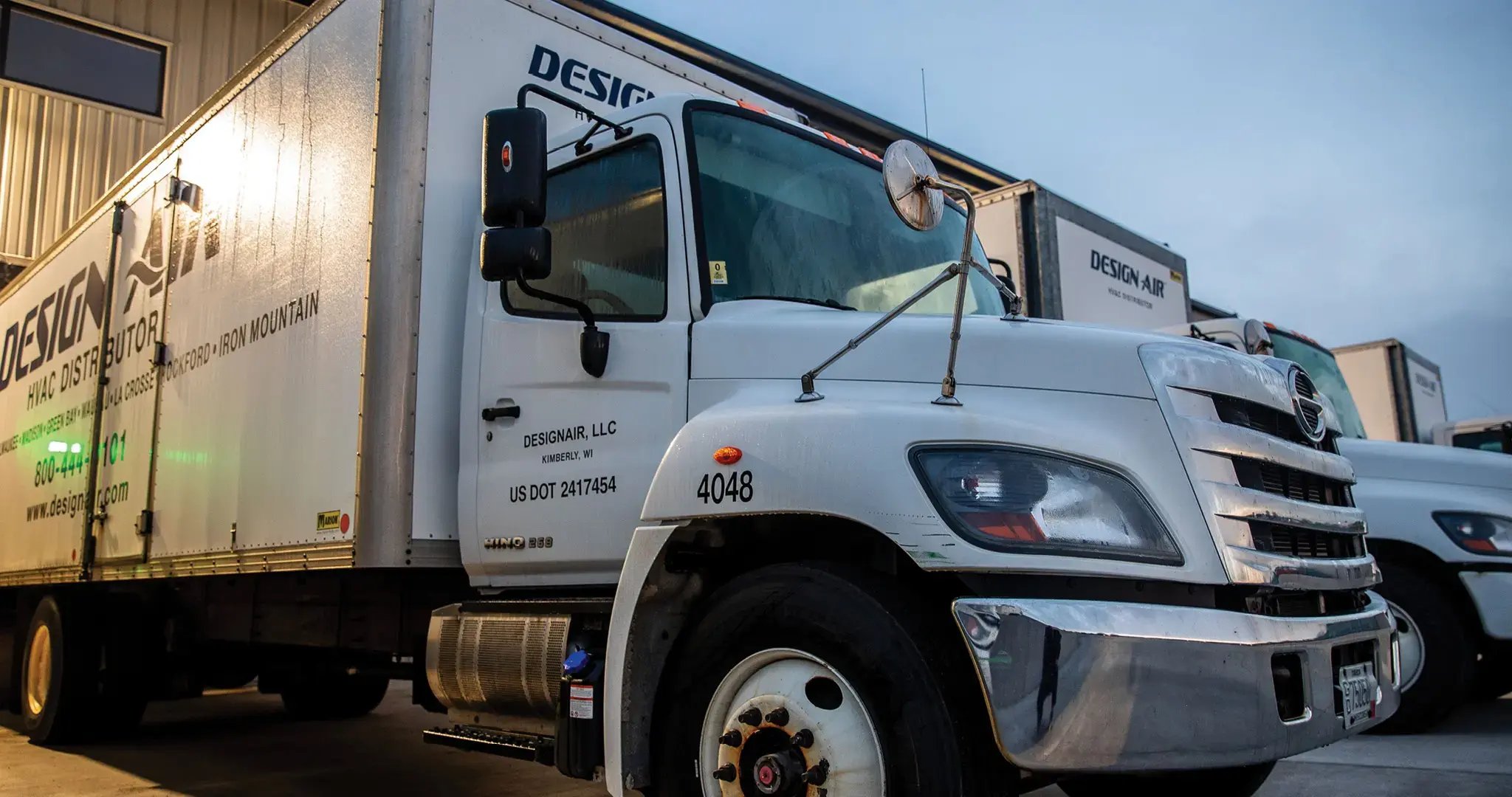 A white Hino Design Air truck parked outside a warehouse on an overcast day and lit from behind by a yellow light