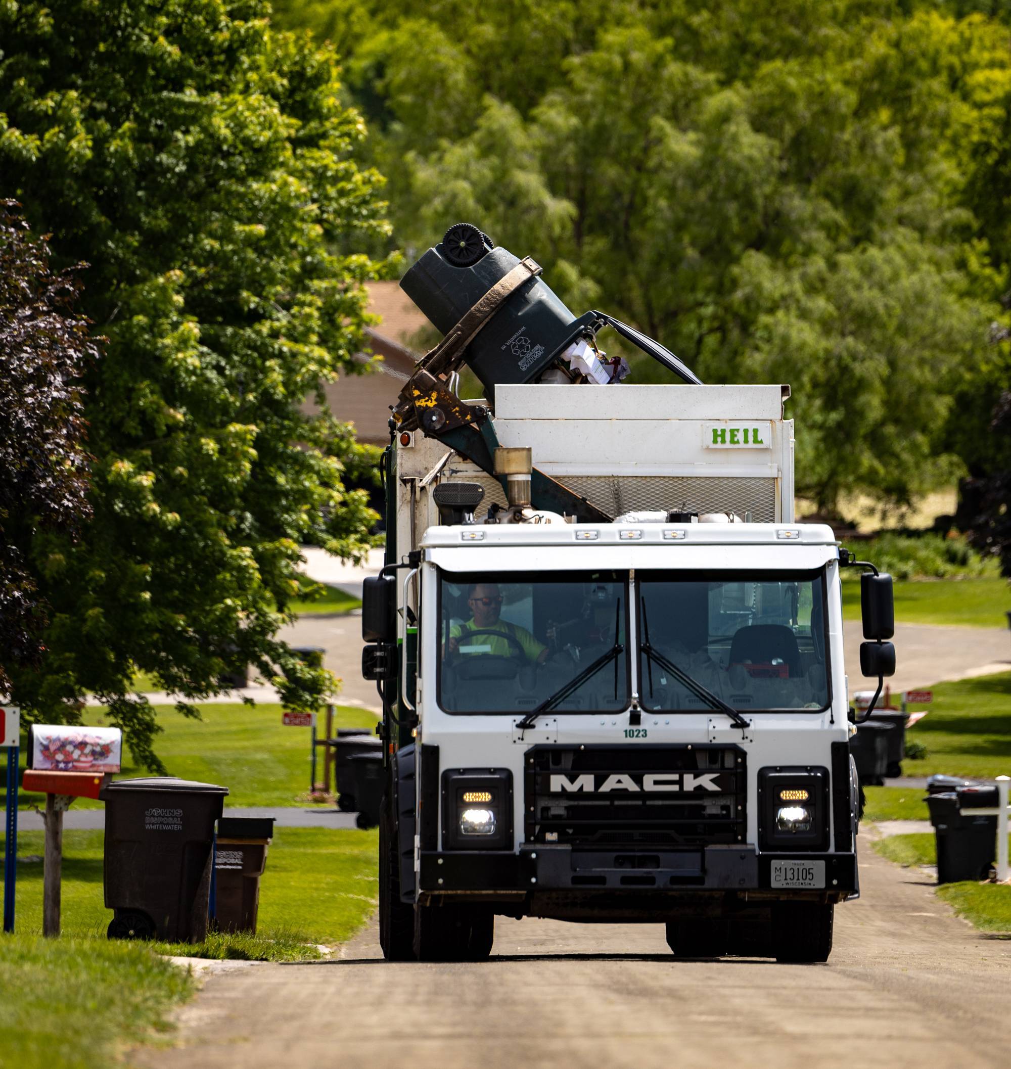 A white Mack truck emptying a garbage can from the front yard of a house in the suburbs