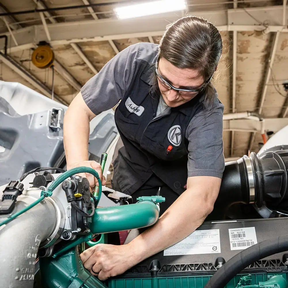 Kriete Technician, Ashley, wearing a dark gray and black overalls and safety goggles, working on a truck engine