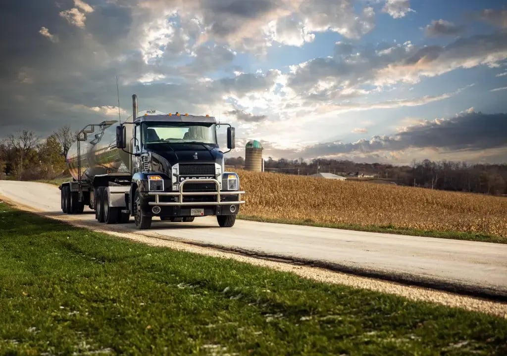 Black Mack truck driving along a country road beneath a cloudy sky, with a field of crops in the background