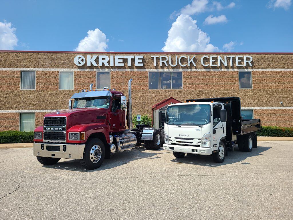 A large red Mack truck and a small white Isuzu truck parked outside a Kriete Truck Center office on a sunny day