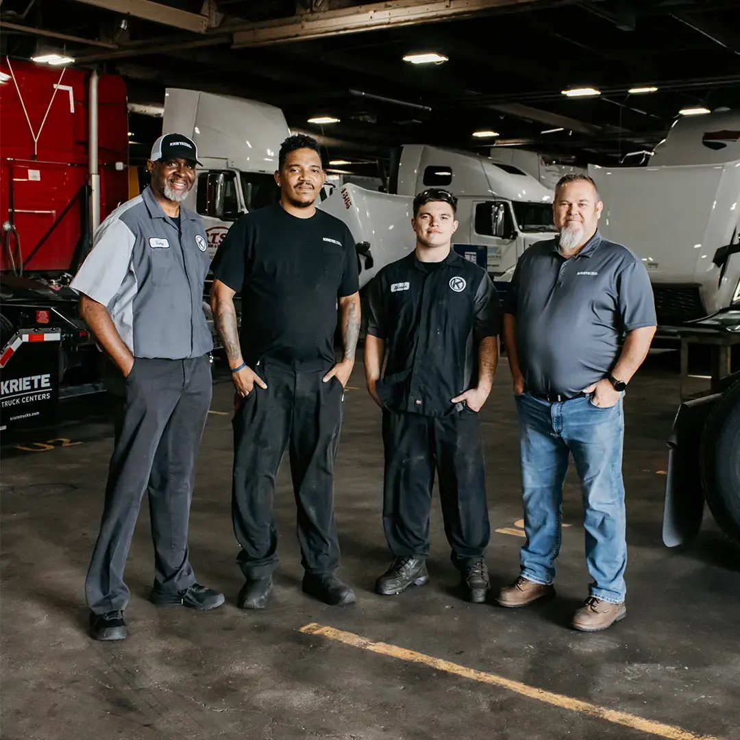 Four Kriete technicians standing together in a dark parking garage with shining trucks in the background